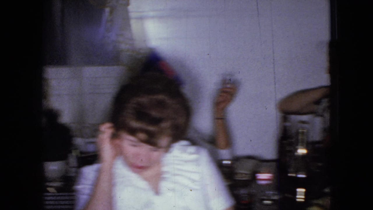 In 1965 home-movie footage from Wellsburg, Iowa, a woman in a white ruffled blouse sits at a table inside a home, with bottles and glasses visible nearby.