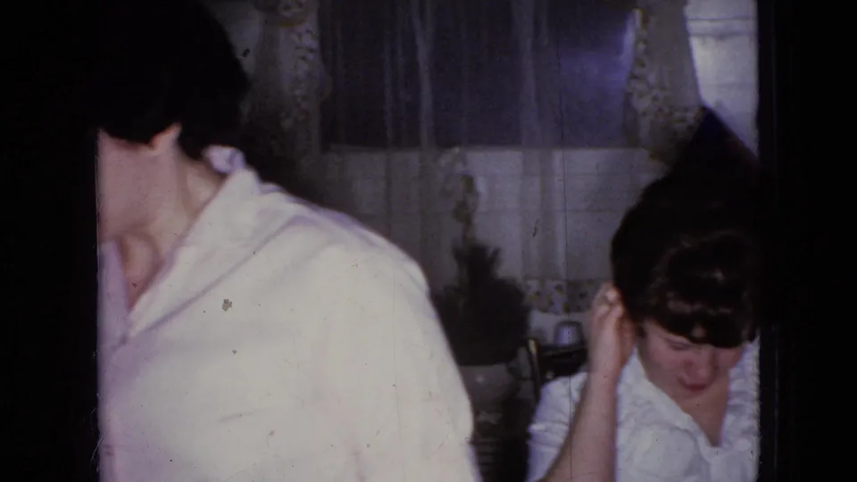 An adult wearing a light-colored shirt turns away from the camera during the Wellsburg, Iowa home dinner scene, with tabletop items and curtains in the background.