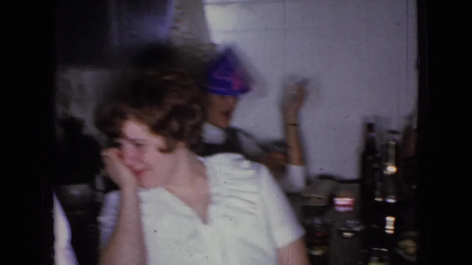 Two adults sit close to a dinner table in the 1965 Wellsburg home-movie scene, while a party hat and bottles suggest an informal celebration or gathering.
