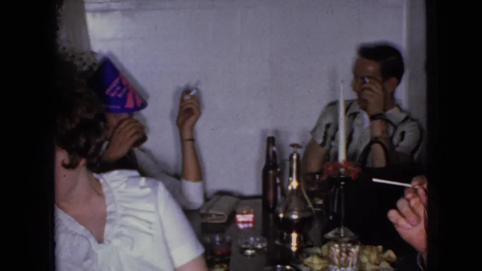 A man in a light shirt smokes a cigarette at the dinner table during the 1965 Wellsburg, Iowa home-movie gathering, with bottles, glasses, food, and other adults nearby.