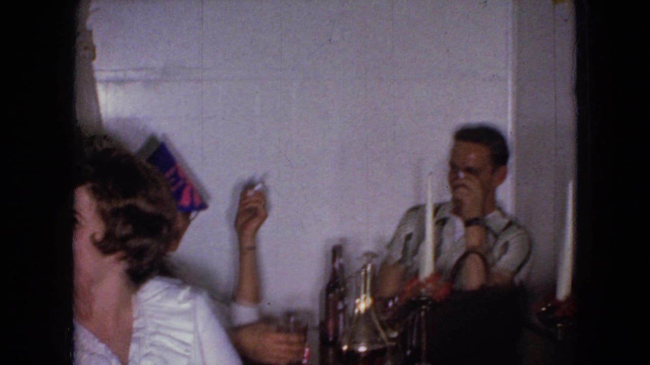 A woman in a white blouse and a man sit near candles, bottles, glasses, and a party hat.