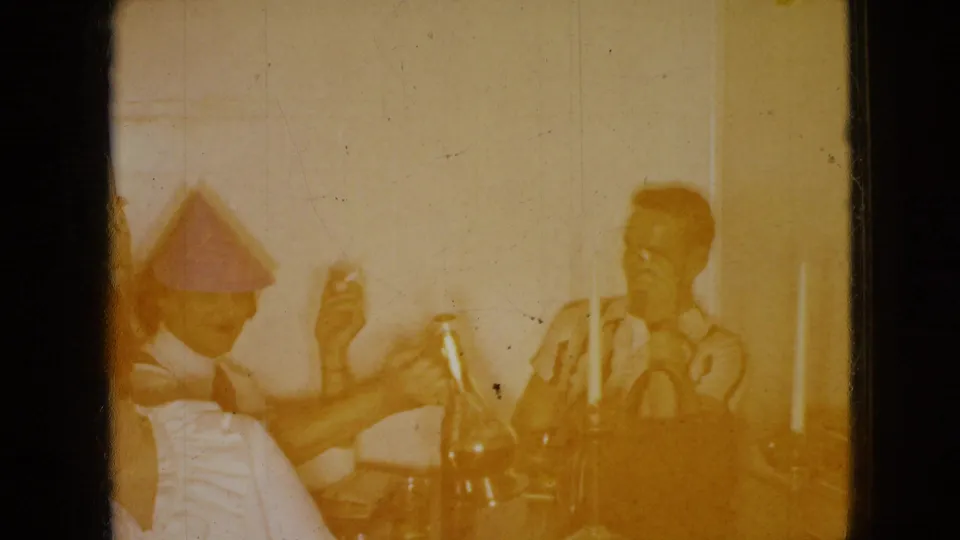 A warm yellow 8mm frame shows adults at the same Wellsburg table with party hats, bottles, candles, and raised hands during a casual gathering.