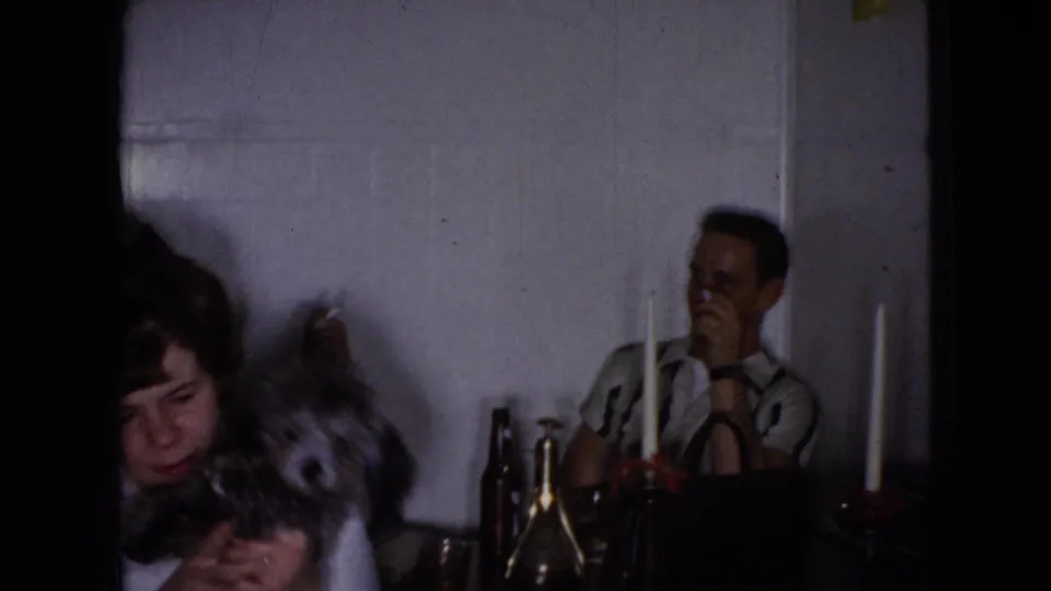 A woman leans toward a light-colored dog beside the table as a man remains seated near bottles, glasses, and candles in the private 1965 home-movie scene.