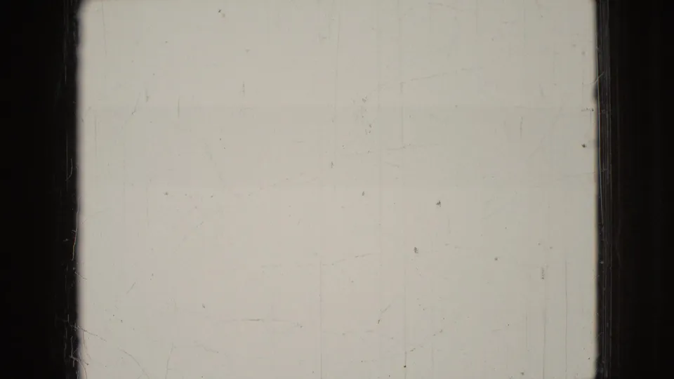 A damaged or transitional 8mm frame from the 1965 Wellsburg, Iowa home movie shows a pale exposed area, dark film borders, and a partial dim view of two dogs and seated adults at the right.