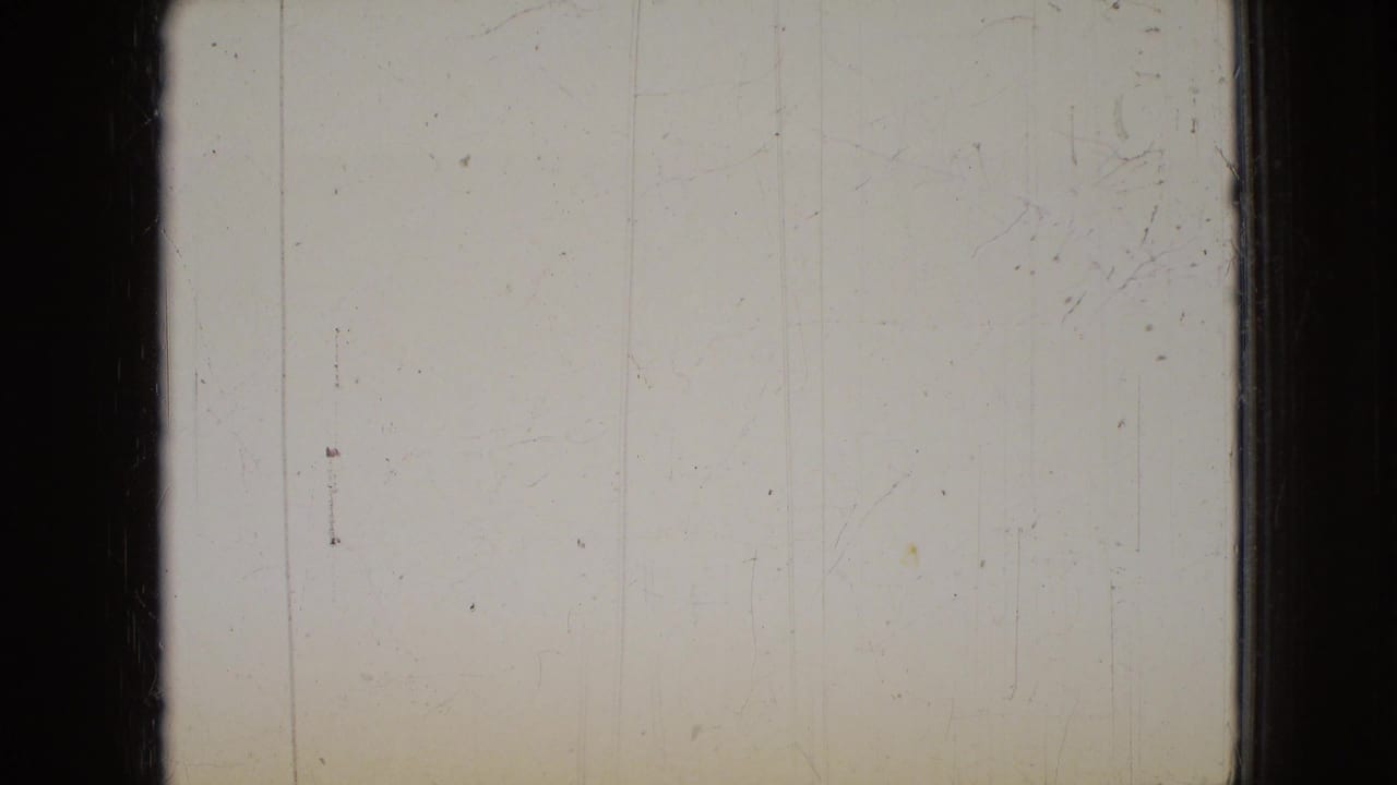 A mostly blank or overexposed 8mm frame with dark film borders, faint vertical marks, and visible surface wear before the living-room action appears.