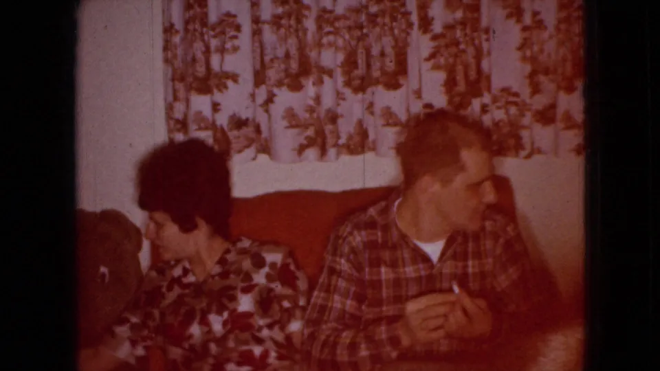 A woman in a floral dress and a man in a plaid shirt sit on a red couch in a private living room, with the man holding a cigarette and both turned slightly off-camera.