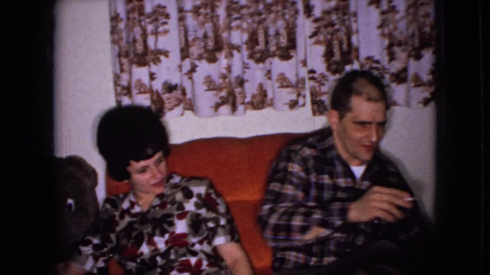 The seated woman looks toward the man in a plaid shirt as he holds a cigarette on a red couch, with patterned curtains and 8mm film borders visible.