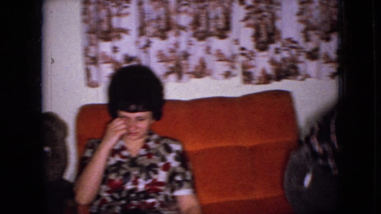 In this 1965 Wellsburg, Iowa home-movie frame, a woman in a floral dress sits on an orange couch beside a large teddy bear with patterned curtains behind her.