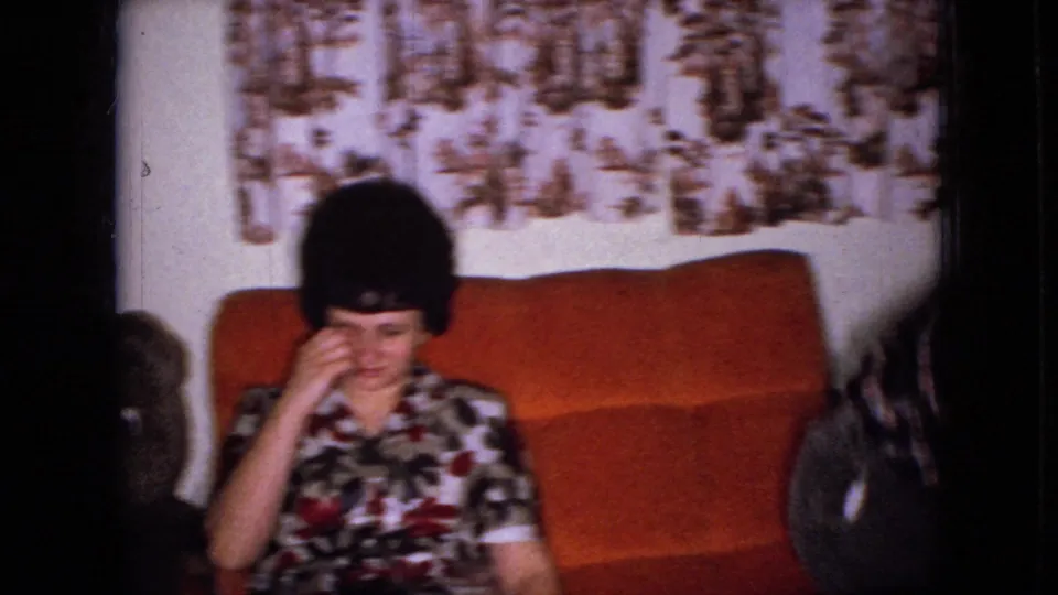 In this 1965 Wellsburg, Iowa home-movie frame, a woman in a floral dress sits on an orange couch beside a large teddy bear with patterned curtains behind her.