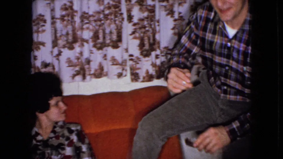The woman seated on the orange couch looks toward a partly visible man in a plaid shirt, with patterned curtains filling the background of the private interior.