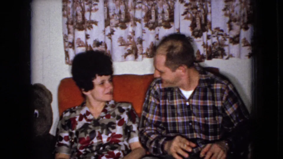 A woman in a floral dress and a man in a plaid shirt sit together on an orange couch in a 1965 Wellsburg, Iowa living room, appearing to converse beside a large teddy bear.
