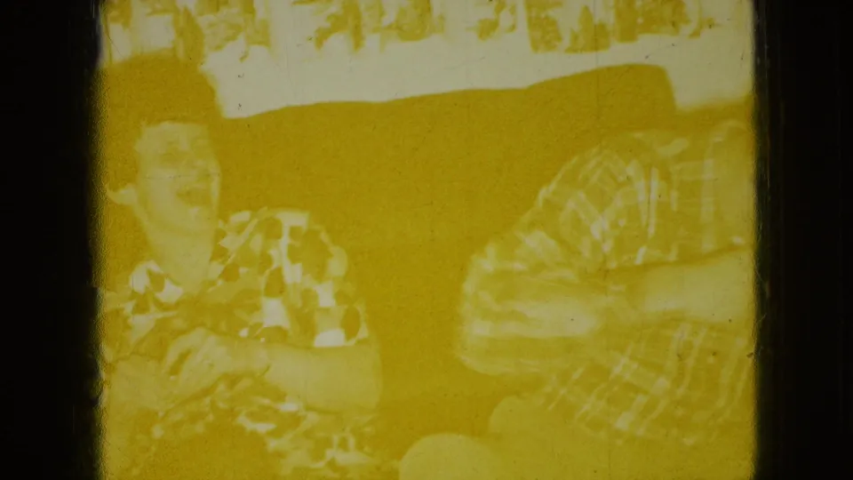 In a 1965 Wellsburg, Iowa living-room scene, a woman in a patterned blouse laughs on a couch beside a man in a plaid shirt, with heavy yellow exposure and frame wear.