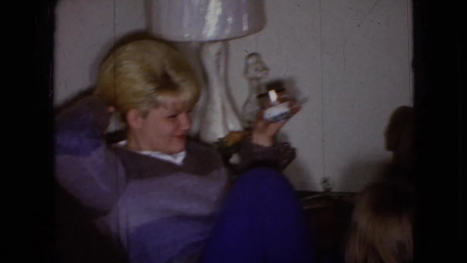 A blond adult reclines in a Wellsburg, Iowa living room near a white-shaded table lamp, holding a small bottle-shaped object against a paneled wall.