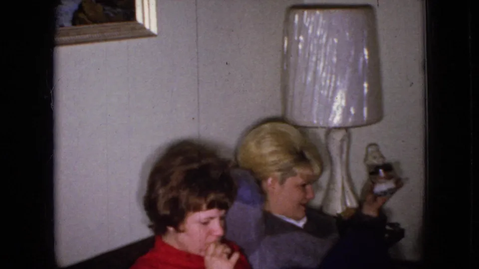 Two adults sit close together in a 1965 Wellsburg, Iowa living room, with a blond person reclining beside a white-shaded lamp and another adult in red in the foreground.