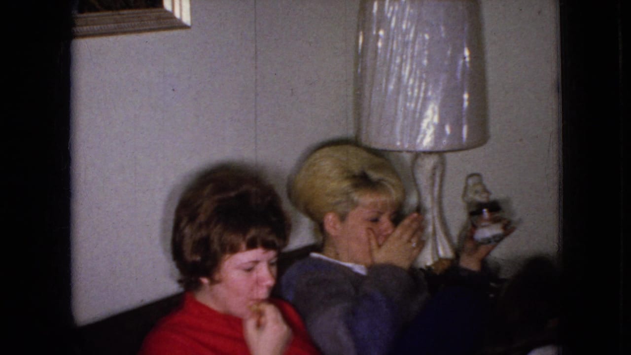 In a 1965 Wellsburg, Iowa home-movie frame, two adult women sit on a couch in a living room beside a lamp, with one eating a snack and the other holding a drink.