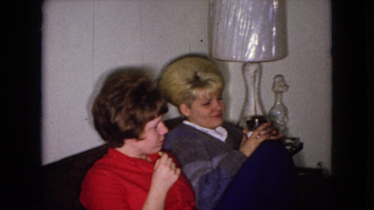 Two women sit side by side, with one looking down and the other holding a glass near a lamp.