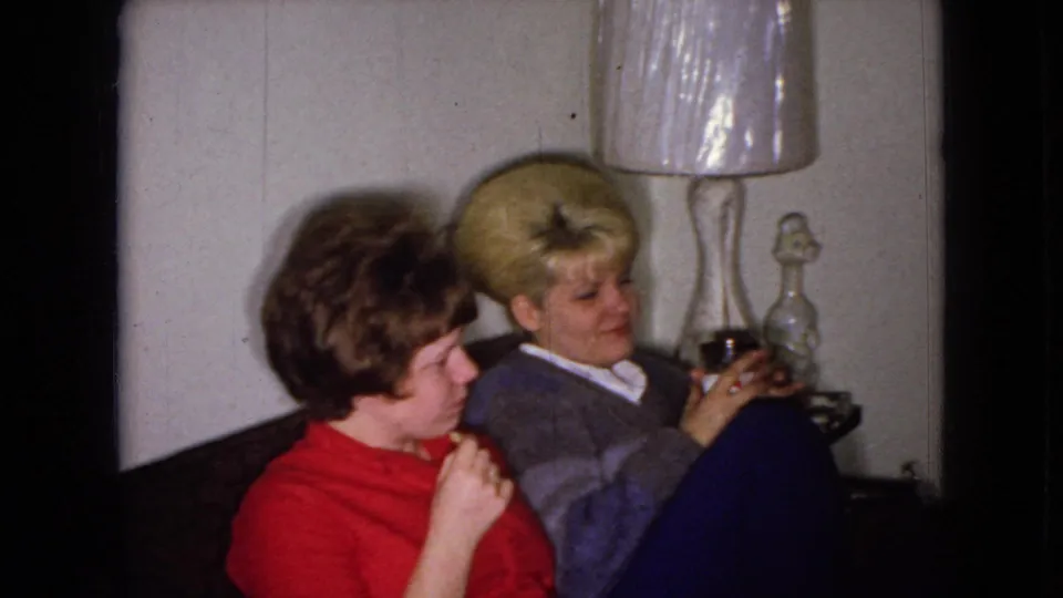In a 1965 Wellsburg, Iowa living room, two adult women sit close together on a couch as the woman with blonde bouffant hair holds a drink near a table lamp.