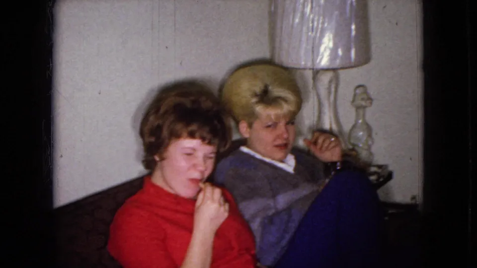A 1965 Wellsburg, Iowa home-movie frame shows two adult women on a living room couch, one eating a snack while the other turns toward the camera beside a shaded lamp.