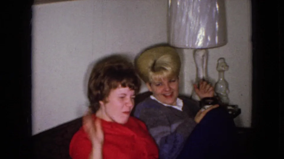 In the closing 1965 Wellsburg, Iowa frame, two adult women seated in a living room smile and wave toward the home-movie camera beside a lamp and tabletop decor.