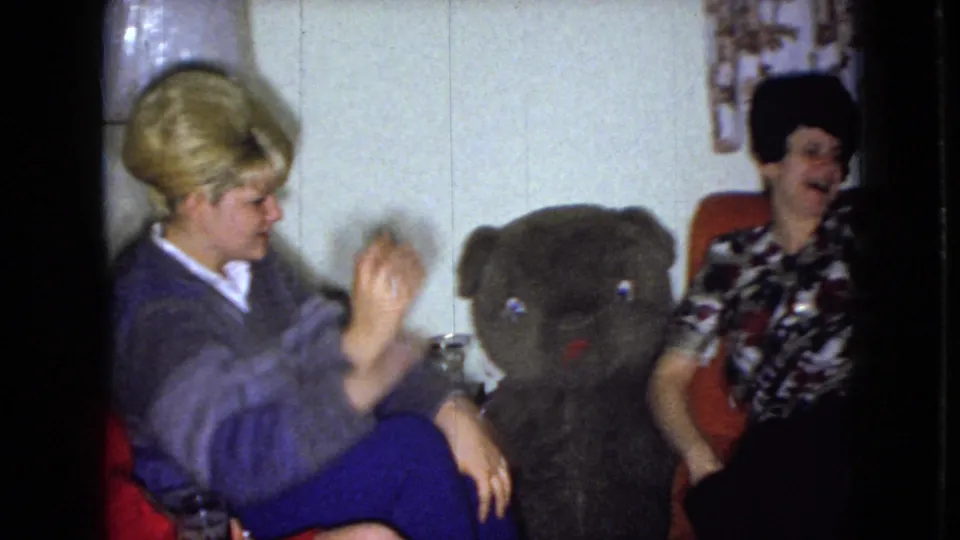 A woman with light hair sits near a large dark teddy bear while another woman in a patterned blouse smiles from the orange couch in the living room.
