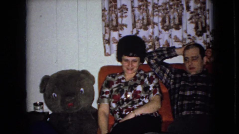 A woman in a floral-patterned shirt and a man in a plaid shirt sit on an orange couch beside a large dark teddy bear, with patterned curtains behind them.