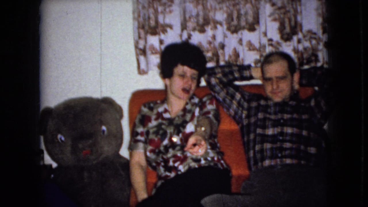 In a 1965 Wellsburg, Iowa home-movie frame, a woman in a floral blouse and a man in a plaid shirt sit on an orange couch beside a large dark teddy bear.
