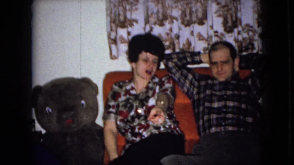 In a 1965 Wellsburg, Iowa home-movie frame, a woman in a floral blouse and a man in a plaid shirt sit on an orange couch beside a large dark teddy bear.