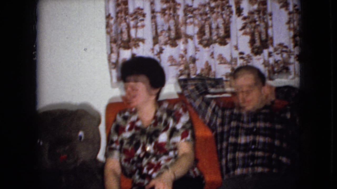 Woman and man sit on a couch with a large teddy bear at the left edge of the room.