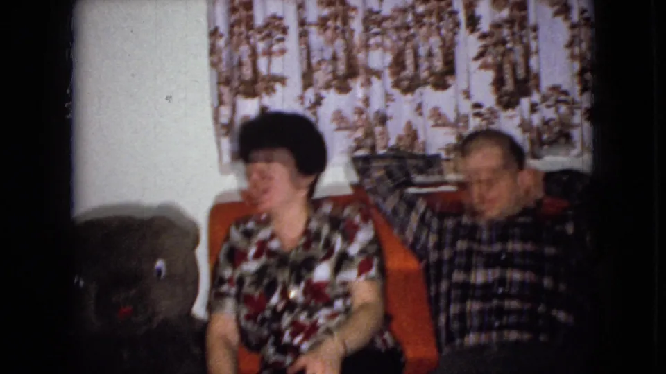 The 1965 home-movie frame shows the seated woman and man from a similar angle, with the teddy bear partly in shadow at the left and patterned curtains behind them.