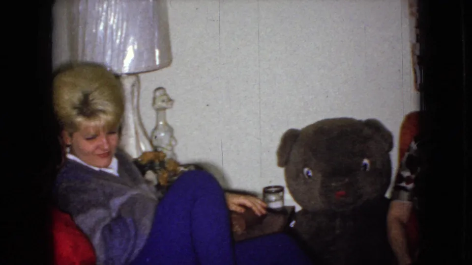 A blond woman in a chair appears near a table lamp and drink, with the large teddy bear centered and part of another seated adult visible at the right edge.