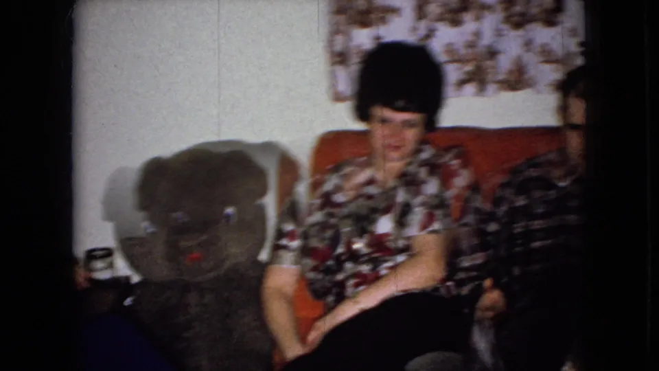 The final 1965 frame holds on the floral-blouse woman seated beside the large teddy bear, with the plaid-shirt man cropped at the right in the private living room.