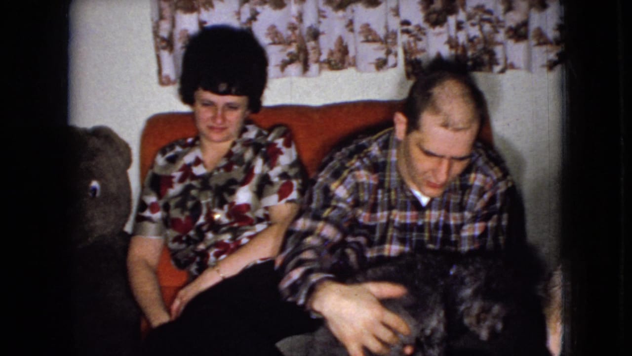 In a 1965 Wellsburg, Iowa home-movie frame, a woman in a floral blouse and a man in a plaid shirt sit on a red couch as he reaches toward a small shaggy dog.