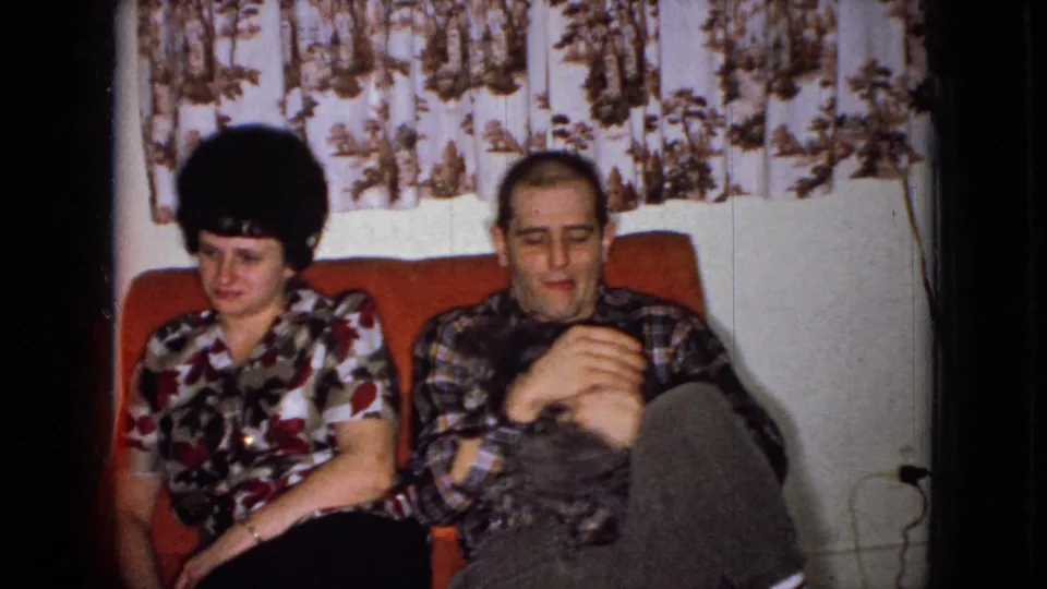 The couple remains seated on the red couch as the small dark dog rests across the man's lap, with patterned curtains behind them.