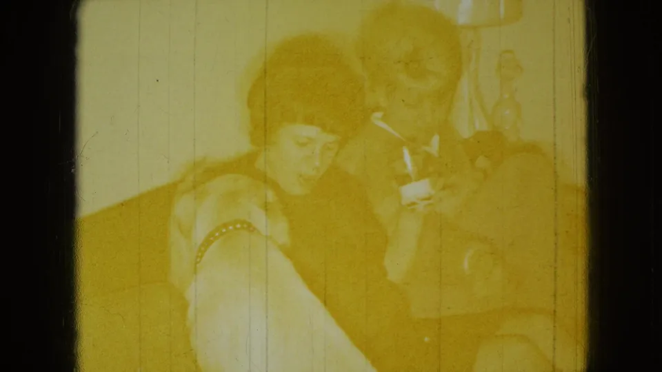 A yellow-faded 8mm frame shows the seated woman and man with a light-colored dog near the red couch, partially obscured by exposure shift and film wear.