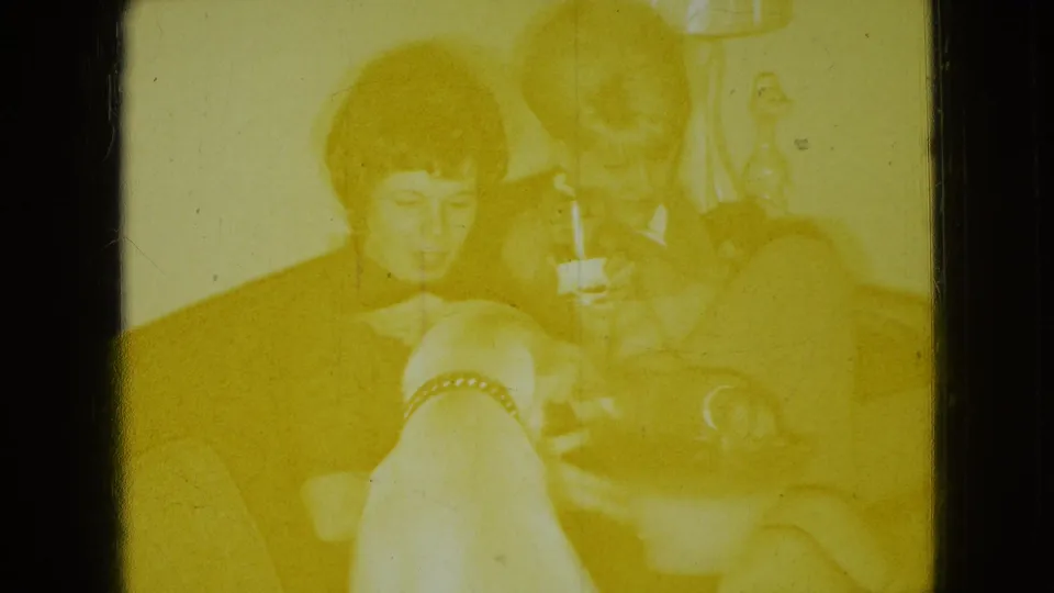 Yellow-shifted 1965 8mm home-movie frame from the Wellsburg, Iowa clip showing adults seated in a living room with a small dog nearby.