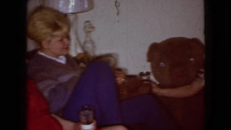 An adult blonde woman sits on the couch in the Wellsburg, Iowa living room, with blue pants, a drink glass, a side lamp, and a large stuffed bear visible nearby.