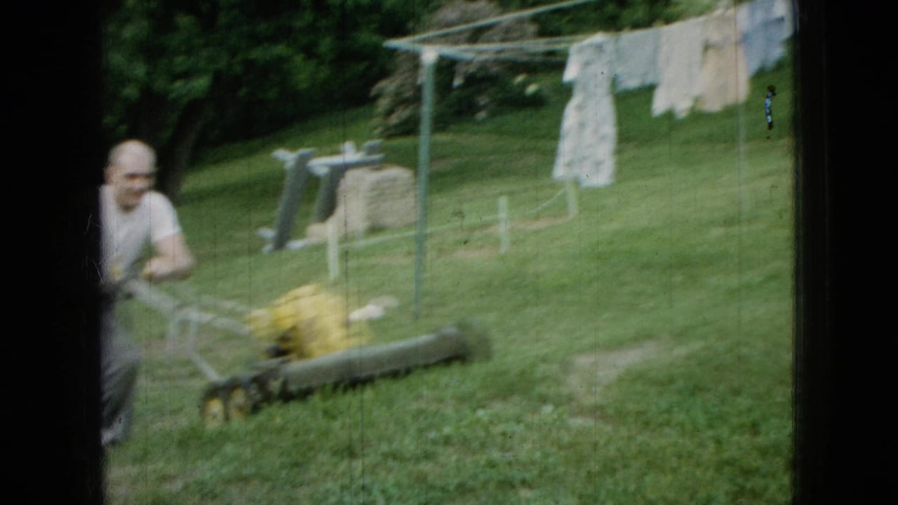 In Wellsburg, Iowa, 1965, an adult man appears partly at frame edge while pushing a yellow mower across a green backyard with laundry on a clothesline behind him.