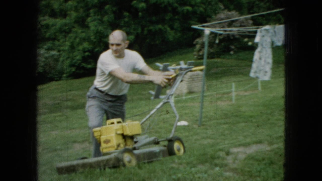 Man in a white T-shirt pushing a yellow mower across grass with laundry behind him.