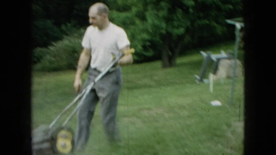 In Wellsburg, Iowa, 1965, the man walks with the push mower across the green backyard, with trees and yard items visible in the background.