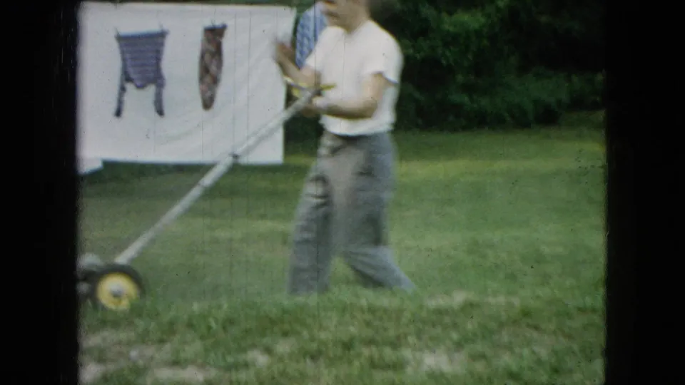 In Wellsburg, Iowa, 1965, the man moves the mower through the yard near hanging laundry, with part of a light-colored outbuilding or wall visible behind him.