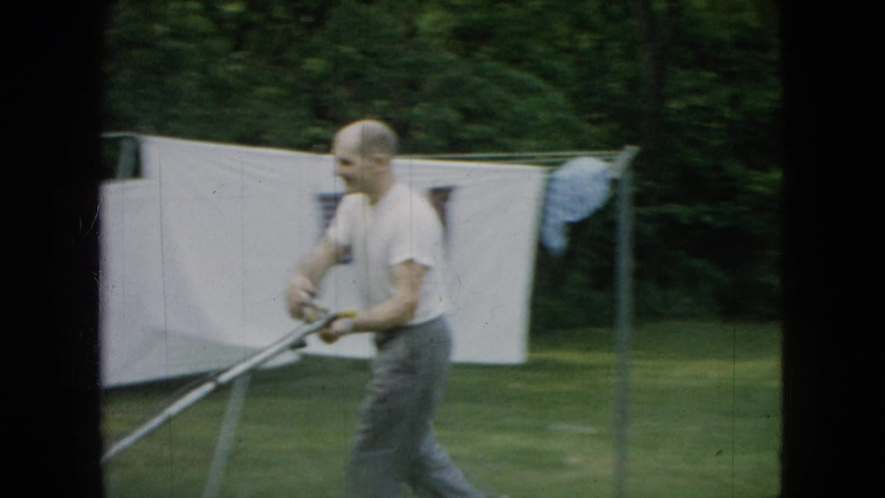 In Wellsburg, Iowa in 1965, a man in a white t-shirt and gray trousers pushes a lawnmower across a green yard near a clothesline and white hanging sheet.