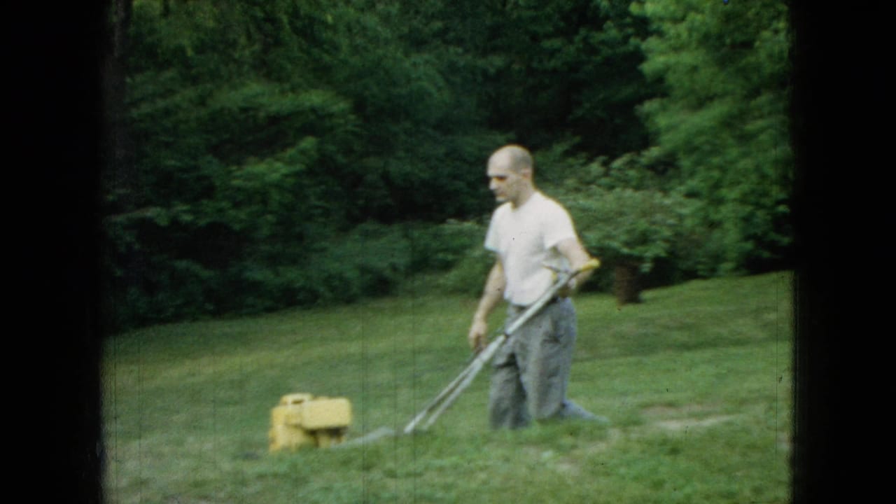 Man walks with a push mower across a lawn with trees and a yellow mower engine visible.