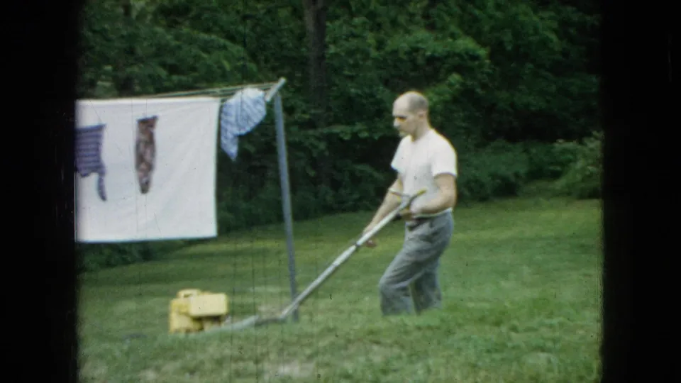 The man continues mowing near a clothesline with a white sheet and garments hanging above the grass, matching the 1965 Wellsburg, Iowa yard-work metadata.