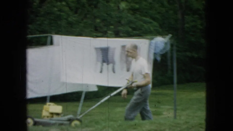 A man in work clothes pushes a vintage lawnmower past a white sheet and hanging laundry in a green yard, with trees in the background of the 8mm home-movie frame.