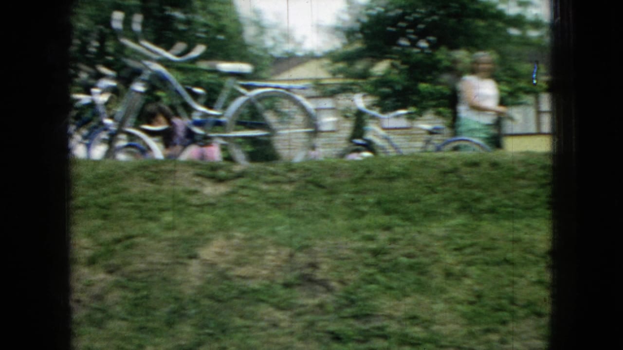 1965 Wellsburg, Iowa home-movie frame showing bicycles near a house, a grassy yard, shrubs, and a blurred person by the outdoor steps.