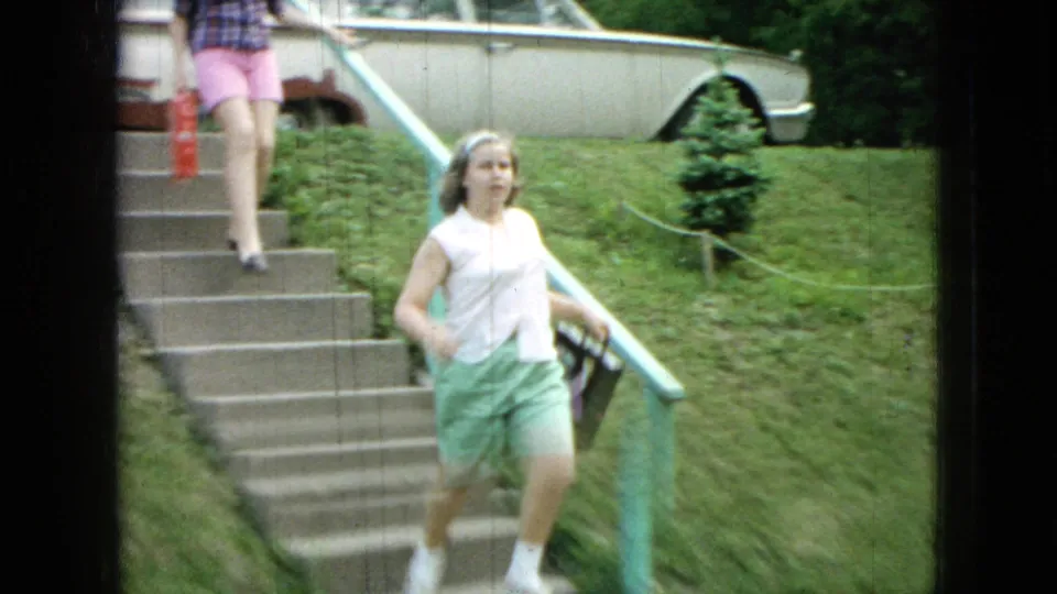 1965 Wellsburg, Iowa frame of a girl in a white sleeveless top and green shorts walking down concrete steps beside a lawn, with another person partly visible behind her.
