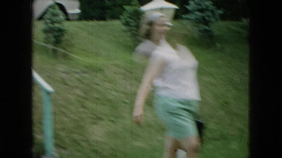 A blurred 1965 Wellsburg, Iowa home-movie frame shows the girl moving past the camera near the steps and lawn, with shrubs and yard details behind her.