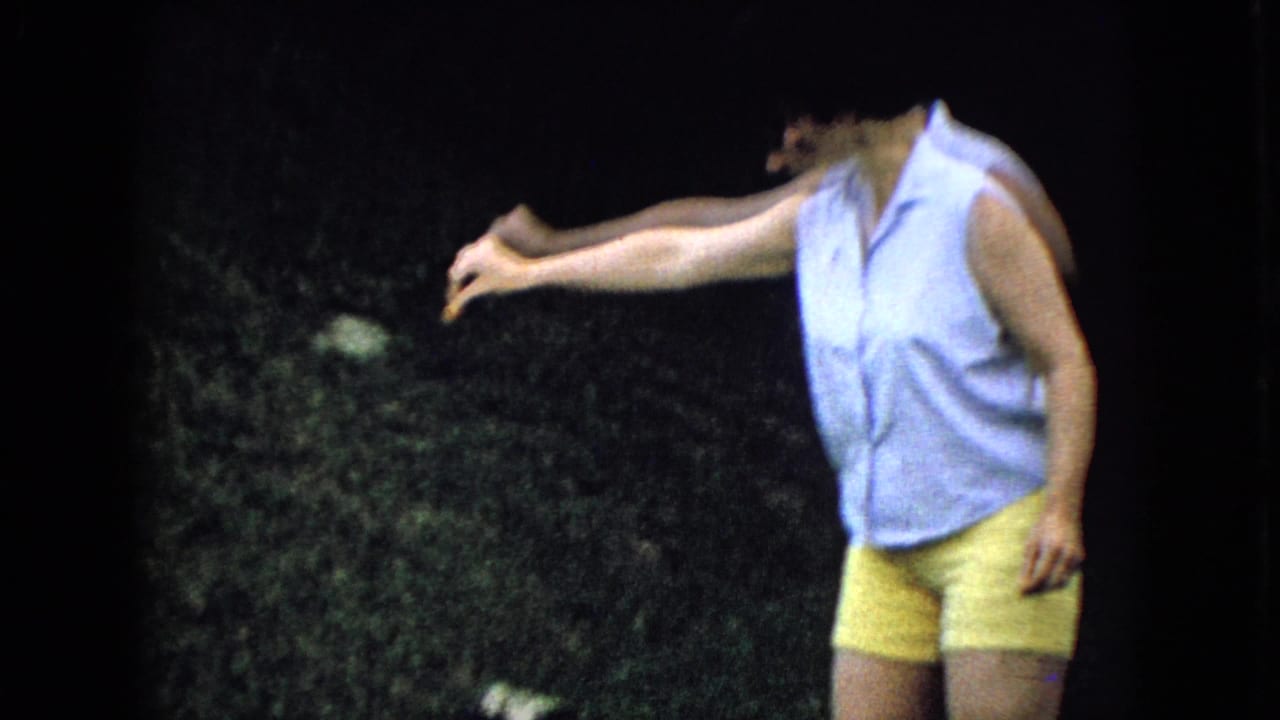 In this 1965 Wellsburg, Iowa home-movie frame, a woman in a light blue sleeveless shirt and yellow shorts reaches one arm outward in a grassy yard.