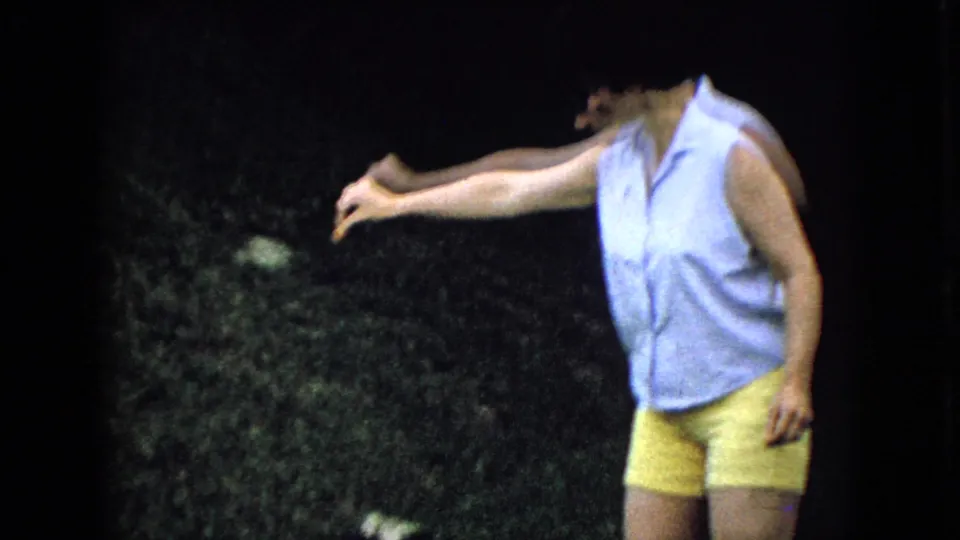 In this 1965 Wellsburg, Iowa home-movie frame, a woman in a light blue sleeveless shirt and yellow shorts reaches one arm outward in a grassy yard.