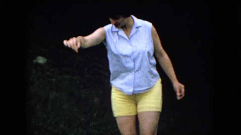 The woman stands in the grassy yard with one hand extended downward, appearing to cue or tease the pug during the outdoor 1965 home-movie moment.